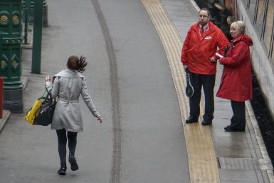 A woman in a trench jacket holding large handbags runs in a British translation with attendants nearby.