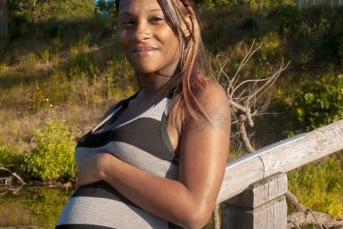 A Black woman in a striped dress smiles standing outside with her hand on her pregnant belly
