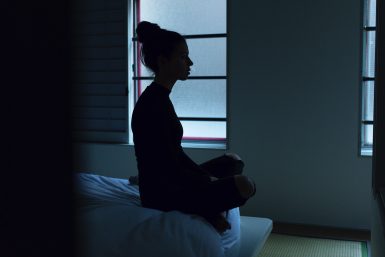 Silhouette of a woman looking to the right, sitting at the edge of her bed in a dimly lit room.