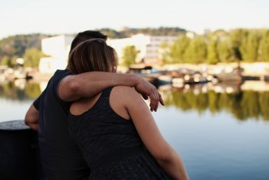 Back view of man with his arm around a woman, looking at a lake outlook.