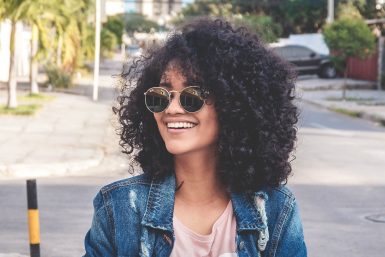 A smiling woman of color, standing on the street in a jeans jacket, pale pink t-shirt, and sunglasses.